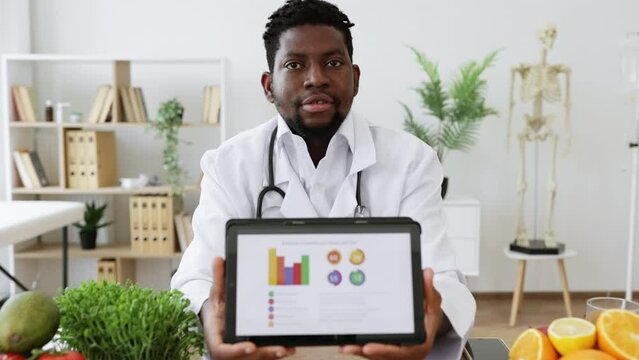 Confident african american male in doctor's coat showing tablet with diet plan on screen while working at hospital. Positive dietitian planning nutrition program on client's personal preferences.