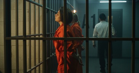 Two female prisoners, inmates in orange uniforms stand facing the metal bars in front of prison cells. Prison officer walks, watches women criminals in jail. Detention center or correctional facility.