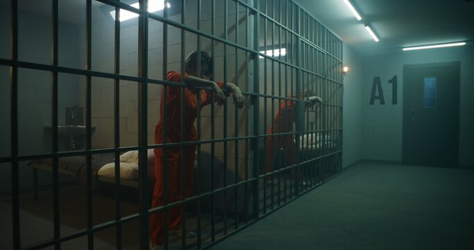 One female prisoner in orange uniform stands behind metal bars, another sits on the bed in prison cell. Women serve imprisonment terms for crimes in jail. Depressed inmates in correctional facility.