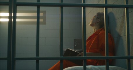 Female prisoner in orange uniform sits on bed behind metal bars, reads Bible in prison cell, looks...
