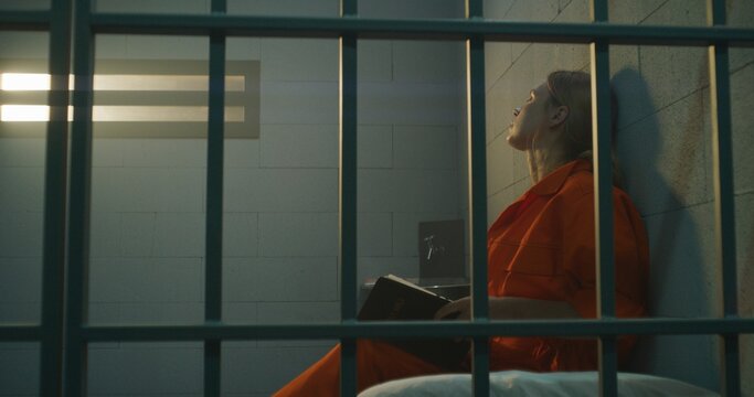 Female prisoner in orange uniform sits on bed behind metal bars, reads Bible in prison cell, looks at barred window. Woman criminal serves imprisonment term for crime in jail or correctional facility.