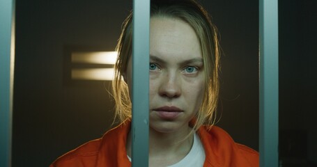 Scared female prisoner in orange uniform holds metal bars, stands in prison cell in handcuffs,...