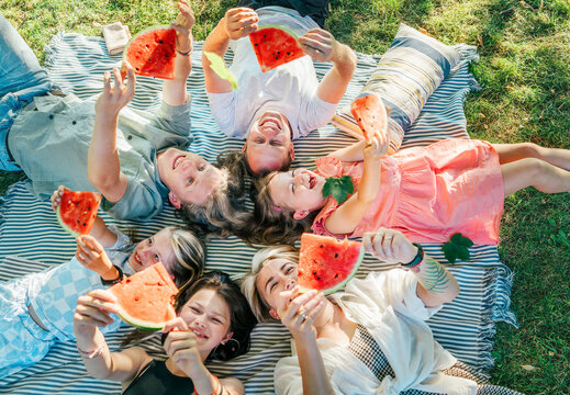 Young daughters with parents family lying on picnic blanket during weekend sunny day, smiling, laughing and eating red juicy watermelon pieces. Family values, fruits vitamins, outdoor time concept