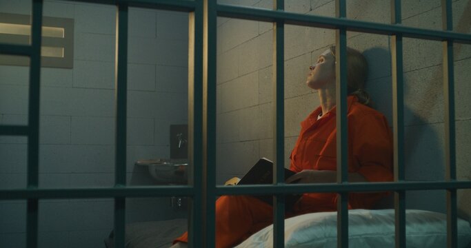Female prisoner in orange uniform sits on bed behind metal bars, reads Bible in prison cell, looks at barred window. Woman criminal serves imprisonment term for crime in jail or correctional facility.