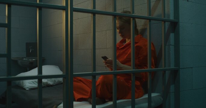 Female prisoner using smartphone in prison. Woman criminal in orange uniform makes call, sits on bed in prison cell. Serving imprisonment term in jail. Detention center or correctional facility.