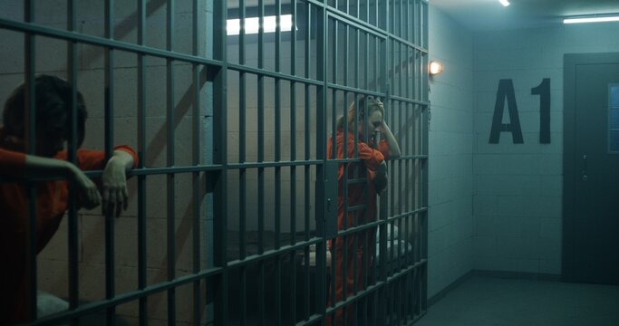 One female prisoner in orange uniform stands behind metal bars, another sits on the bed in prison cell. Women serve imprisonment terms for crimes in jail. Depressed inmates in detention center.