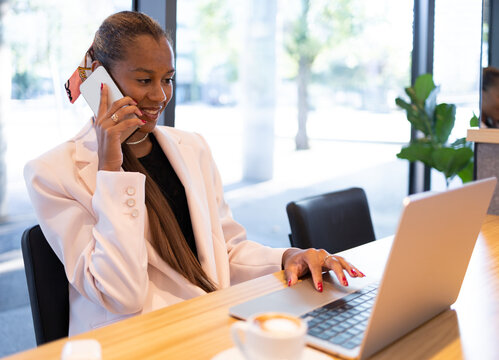 Cheerful ethic woman talking on smartphone while working via laptop