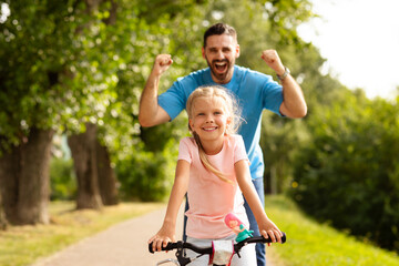 Happy father teaching daughter to ride a bike in park, dad shaking fists on background, summer fun...
