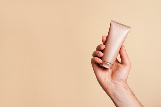 Male hand holding unbranded beige tube with lotion or cosmetics cream on yellow background. Shaving foam or male skin care concept. Minimalism, copy space