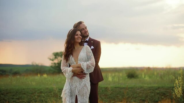 Just meried couple standing in the field with flowers at sunset. The husband embraces his wife from behind, and they cuddle up to each other. Incredibly beautiful sky In the background.