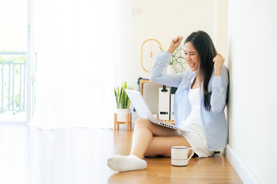 Young Asian beautiful woman working at home, lady in confortable out fit using laptop on her lap, rising her two hand with smile and luagh with happiness and copy space.