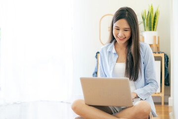 Young Asian beautiful woman sitting on floor working with her laptop at home with smile of happiness at living roon, glass door and white curtain with copy space