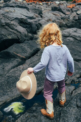 girl with her back turned climbing on the rocks. by the ocean with a hat in her hand. 