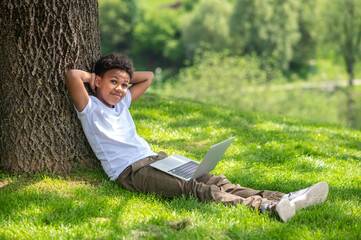 Cute boy sitting in park under tree using laptop to communication with friends or online education.