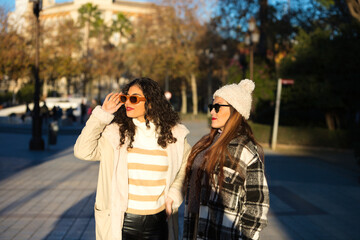 Obraz premium two young, beautiful, Spanish women who are friends dressed in winter clothes and wearing sunglasses are walking and enjoying the sunny day in seville, spain.