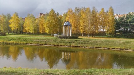 In the city park, a rotunda of concrete columns with a metal roof in the form of a dome, surrounded by shrubbery, stands next to birch trees yellowing in the fall and a water canal