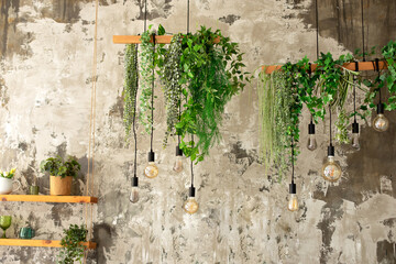 Many different retro Edison light bulbs with garland from green plants and leafs hanging from ceiling. Loft pendant lamps on background of rough cement plaster on wall. Vintage Edison light bulbs.	
