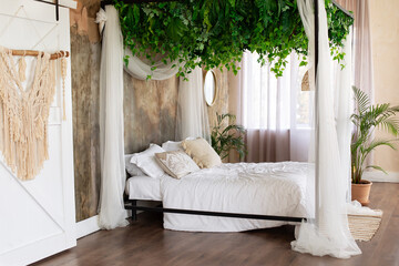 Cozy light bedroom in Scandinavian style bedroom, canopy bed, plant in pot. Bed decorated flowing white curtains canopies. Interior boho design bedroom with composition from garland flowers and plants