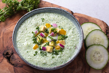Green cucumber gazpacho cold cream soup styled and decorated in rustic bowl with ingredients on gray table, top view
