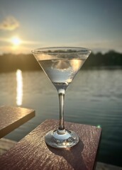A martini cocktail sits on the arm of a red chair by a sunset over the water.