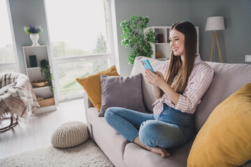 Photo of sweet cute young girl wear striped shirt sitting sofa texting messages modern gadget indoors apartment room