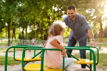 Fototapeta premium Child girl having fun with her father on outdoor playground, dad riding daughter on carousel, spending time together