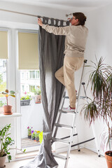 A woman stands on a stepladder and hangs gray curtains on a curtain rod in an apartment, spring...