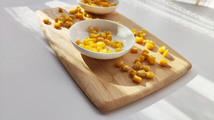 Lot of pieces of canned yellow corn on plate which is on wooden bamboo cutting board on white background. The concept of cooking and delicious healthy food