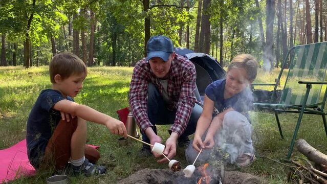 Delightful family camping trip. Father and children gather around campfire to roast marshmallow on wooden sticks in the great outdoors on sunny day. Outdoor cooking while hiking adventure in forest