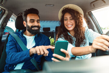 Cheerful arab man showing road on phone to female driver holding and using smartphone, couple looking at digital map app