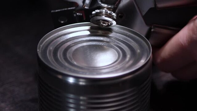 Close-up strong male hands with a can opener make an incision on the lid of a metal can on a black table.