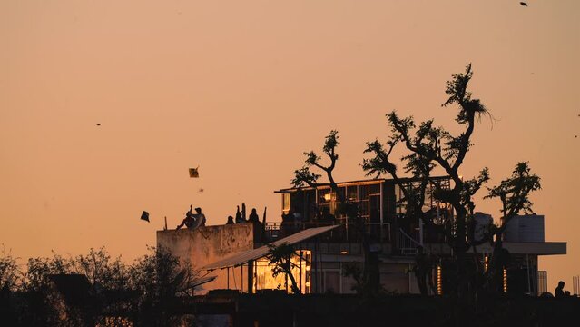 orange dusk shot with silhouette of people on roof of glass building with kites flying in the sky on the traditional festival of sankranti uttarayan independence day in India