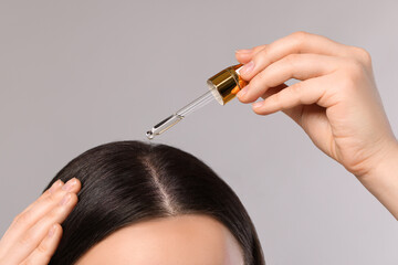 Woman applying essential oil onto hair roots on light grey background, closeup