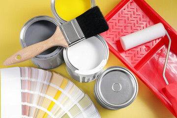 Cans of paints, brush, roller, tray and palette on yellow background, flat lay