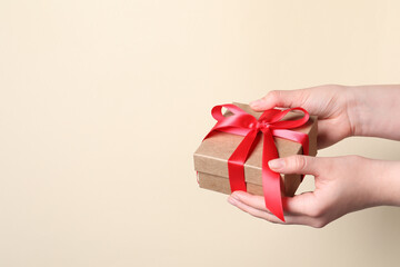 Woman holding gift box with red bow on beige background, closeup. Space for text