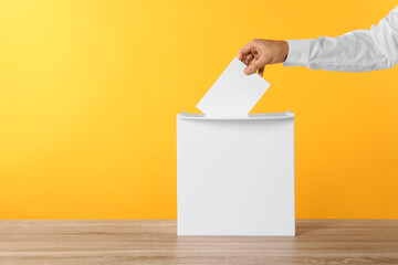 Man putting his vote into ballot box on yellow background, closeup. Space for text