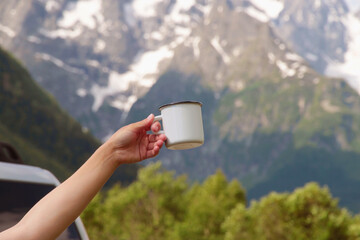 The traveler holds a cup of coffee in the mountains. White mug in the hands on the background of the mountains. Mountain camping concept.