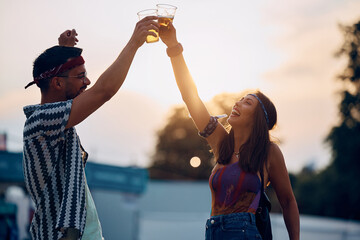 Carefree couple toasting with beer while having fun on summer music festival at sunset.