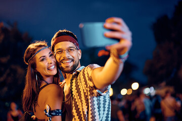 Happy couple of festivalgoers taking selfie during music concert at night.