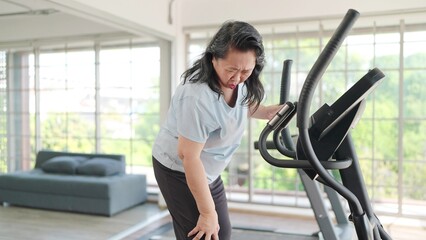 Fototapeta premium Asian elderly women suffering legs or knee pain from exercising on the machine in living room at home. Old age, Health problem and people concept. Pain from exercise