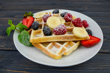 Belgian waffles with summer berries and powdered sugar in a white plate on a dark wooden background.