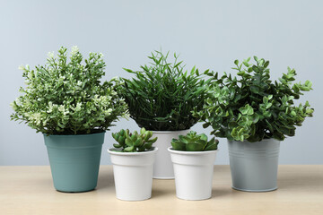 Many different artificial plants in flower pots on wooden table against light grey background