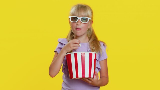 Excited young cute school girl in 3D glasses eating popcorn, watching interesting tv serial, sport game, film, online social media movie content. Teen female child kid on studio yellow background