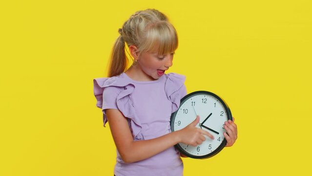 It is your time. Young school girl showing time on wall office clock, ok, thumb up, approve, pointing finger at camera, time for education study. Preteen child kid isolated on studio yellow background