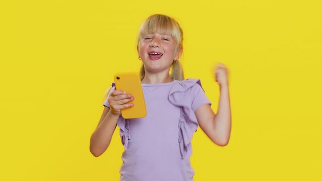Young preteen child girl kid use mobile smartphone typing browsing say Wow yes found out great big win good news celebrate doing winner gesture. Little cute children on studio yellow background