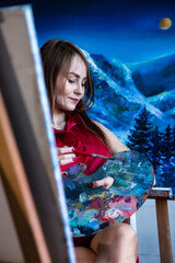 Lovely woman painter in red shirt painting with brush oil paints on canvas in art lab, pensive...