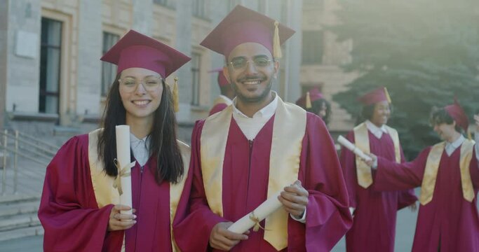 Caucasian woman and Arab man wearing graduation caps and gowns standing on campus holding diploma scrolls and smiling. People and education concept.