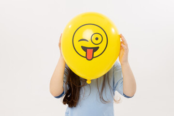 A child girl hides his face behind a yellow winking emoticon on white background. Funny face. World Emoji Day