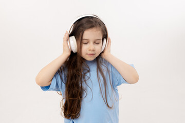 Happy smiling child girl enjoys listens to music in headphones over white background. Vivid and fun emotions, happy child with pleasure listens to songs in wireless headphones