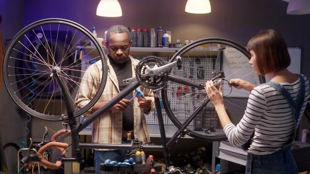 Two professional mechanics talking at workplace. Man and woman fixing bicycle chain at repair shop. Female smiling and working with screwdriver. Focused male and female discussing at workshop.